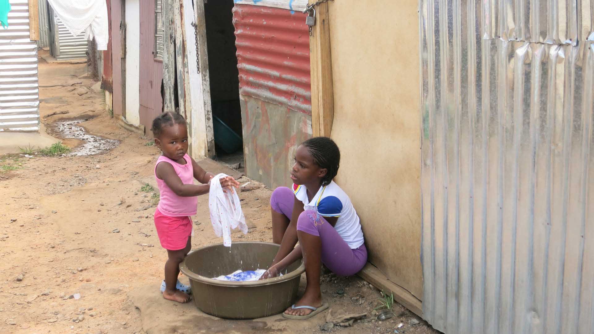 Zwei Kinder waschen Kleidung vor Wellblechhütten auf sandigem Boden in Johannesburg – Kulturbegegnung auf einer Trekkingreise