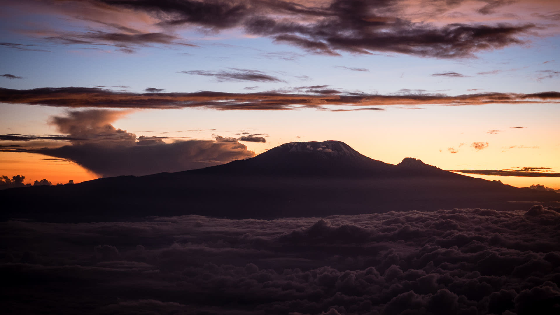 Trekking am Kilimandscharo: Gipfelsilhouette mit Schneeplateau über Wolkenmeer bei orangefarbenem Sonnenaufgang auf der Machame