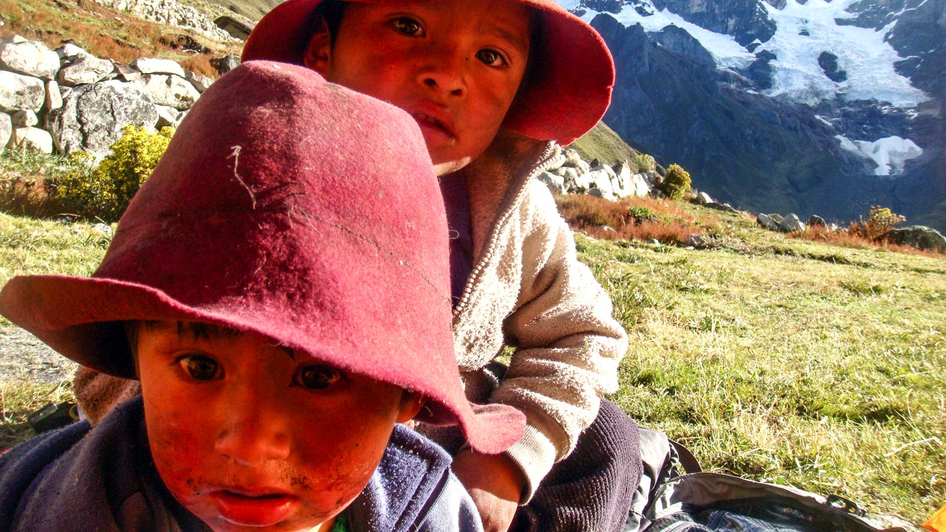 Zwei peruanische Kinder mit roten Filzhüten auf einer Hochebene der Cordillera Huayhuash, Gletscher im Hintergrund.