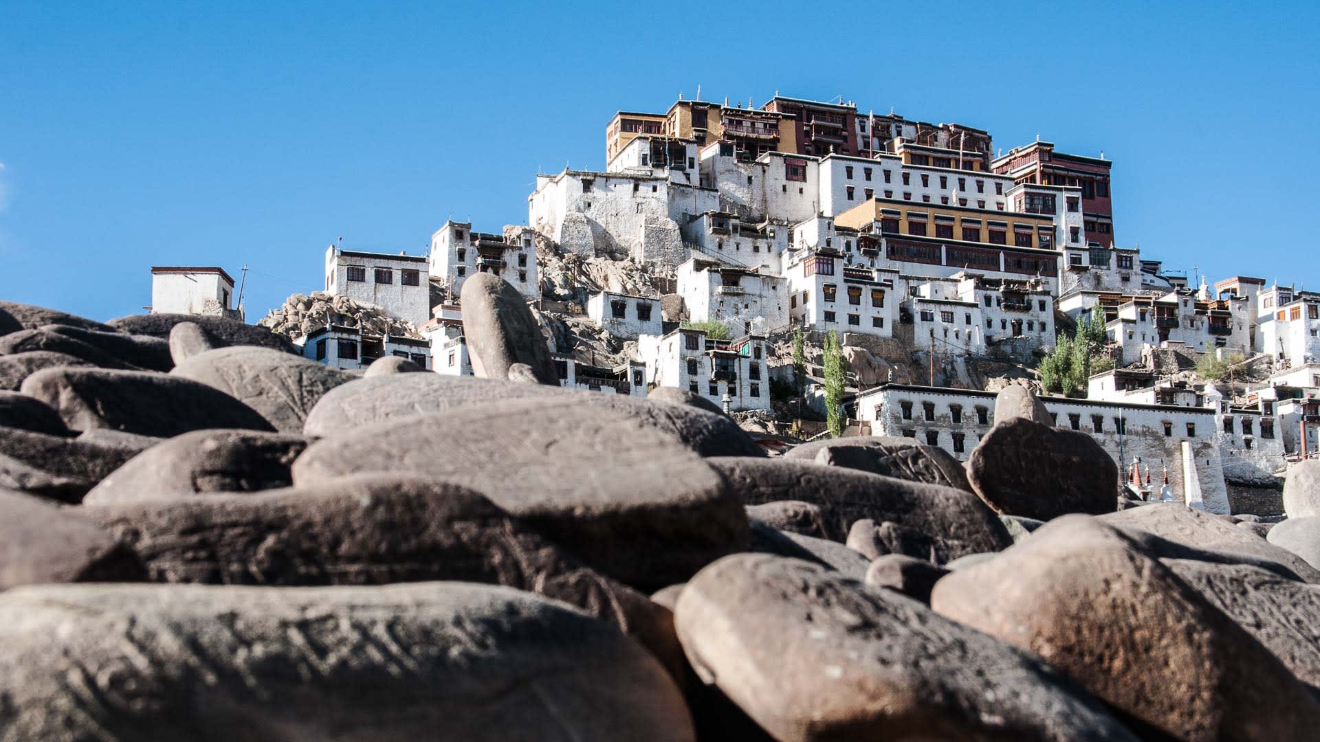 Trekking in Ladakh: mehrstöckige Klosteranlage auf Felsenhügel mit Mani-Steinen im Vordergrund unter tiefblauem Himmel.