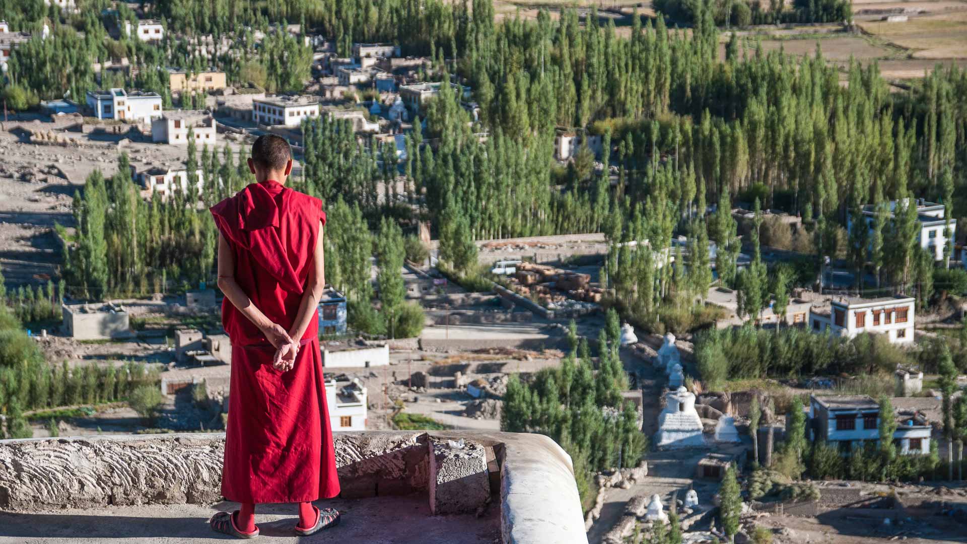 Trekking in Ladakh: Person in rotem Gewand steht auf Steinterrasse und blickt auf eine Talsiedlung mit weißen buddhistischen Stupas