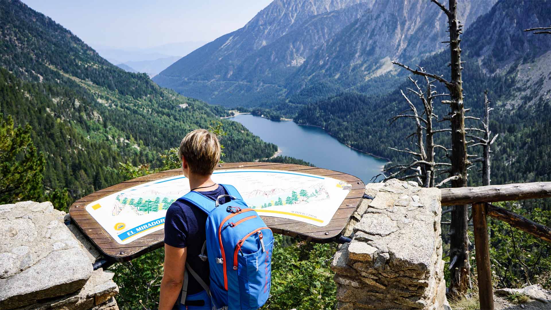 Trekker mit Rucksack am Mirador de Sant Maurici liest Panoramatafel mit Blick auf Bergsee im Tal bei Espot.