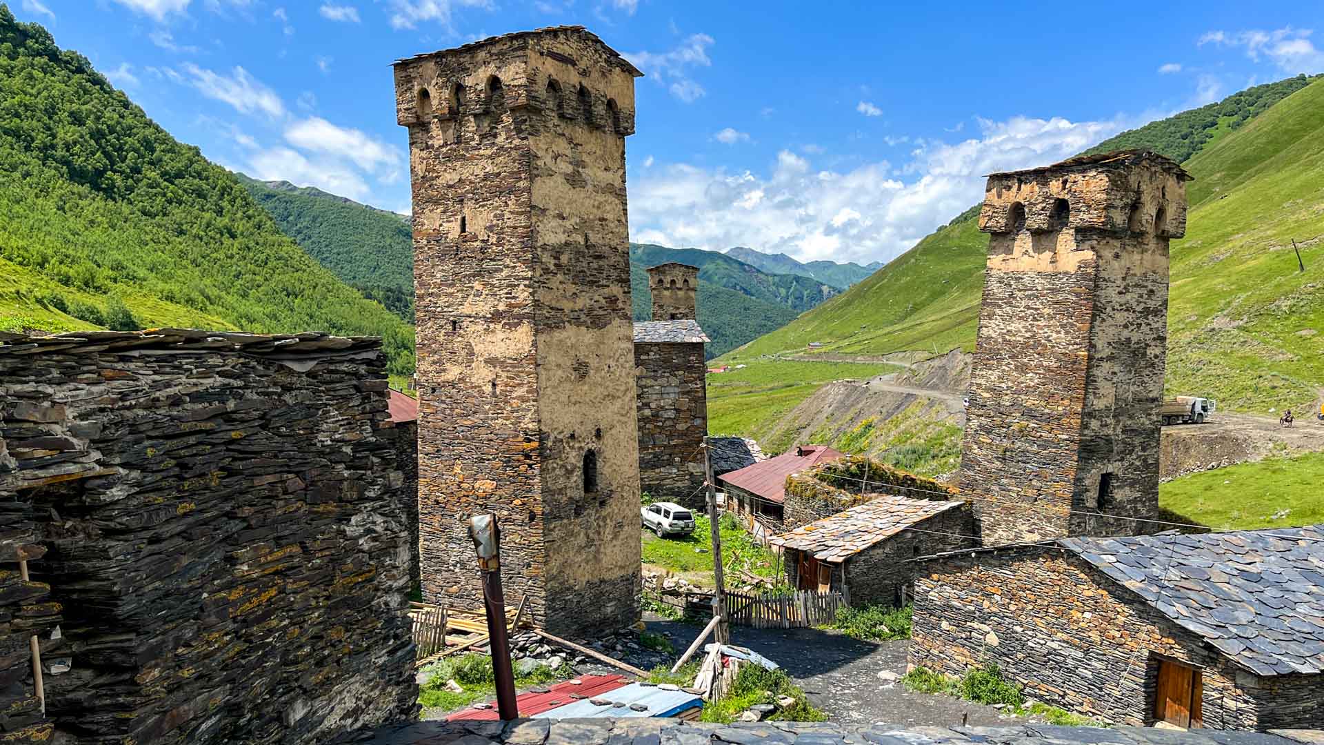 Trekking in Swanetien: Mittelalterliche Steintürme eines Gebirgsdorfs mit Schiefer-Gebäuden und grünen Berghängen im Hintergrund.