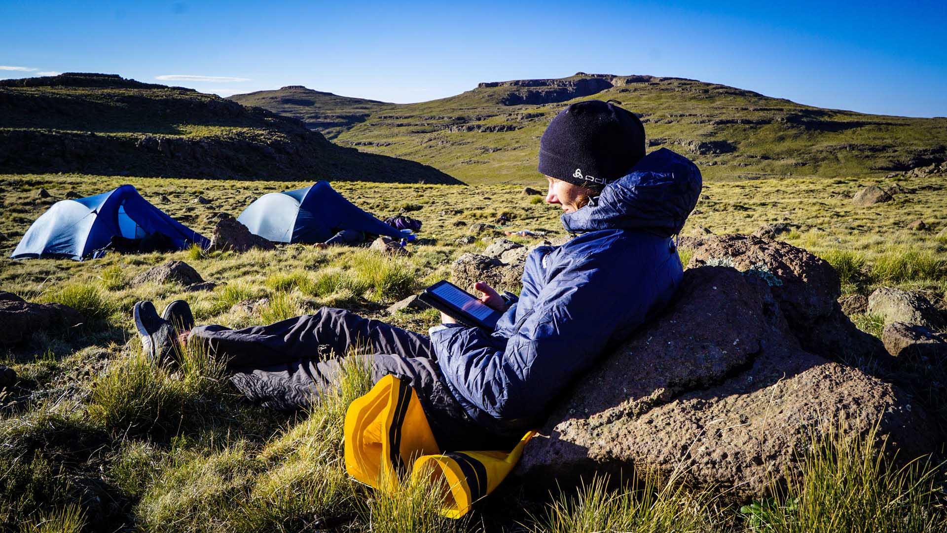 Trekker pausiert angelehnt an Felsblock im Camp auf dem Hochplateau der Drakensberge mit Kuppelzelten.