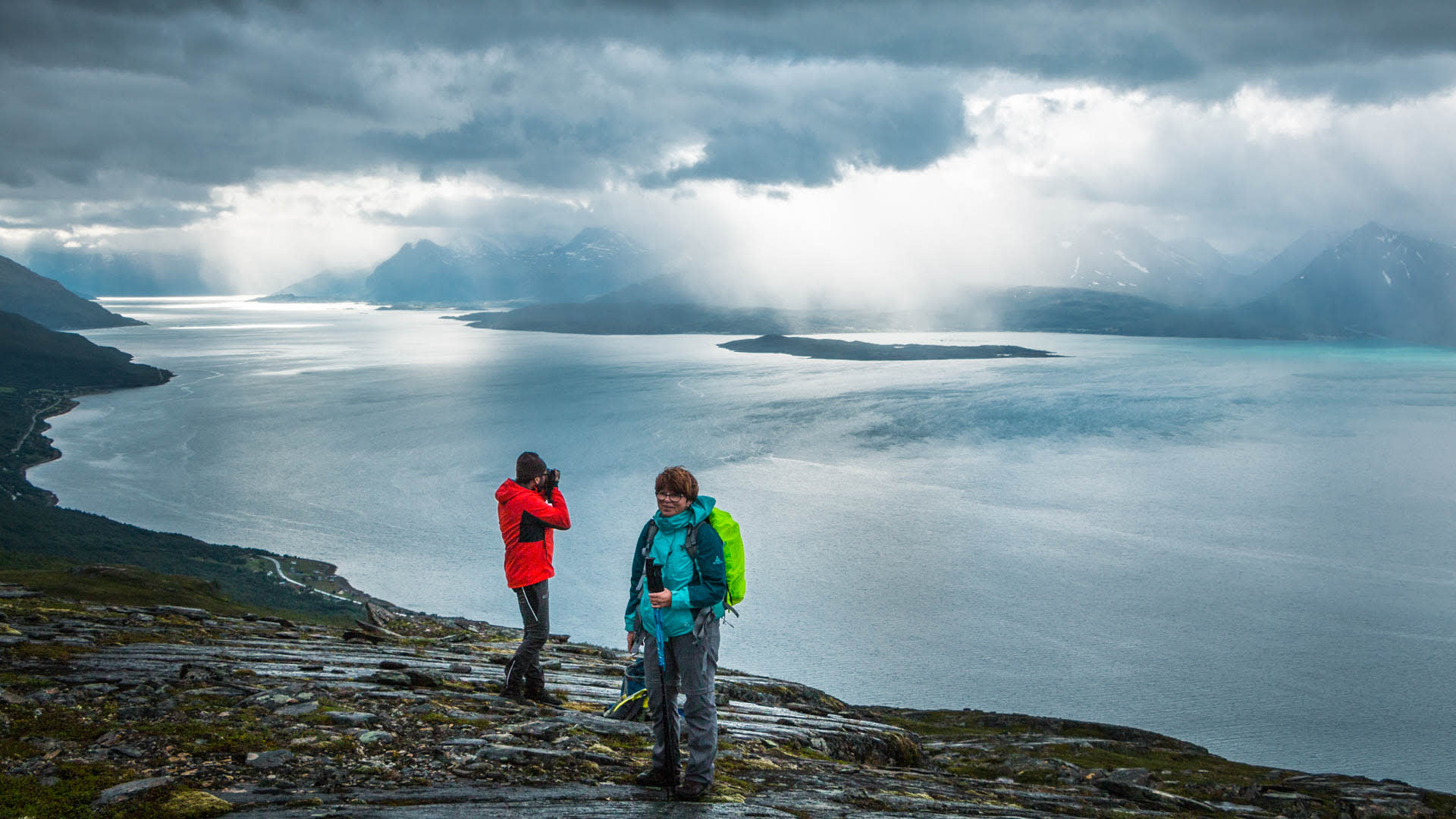 Zwei Trekker auf Felsplatten am Storhaugen mit Blick über den Lyngenfjord unter bewölktem Himmel mit Lichtstrahlen.