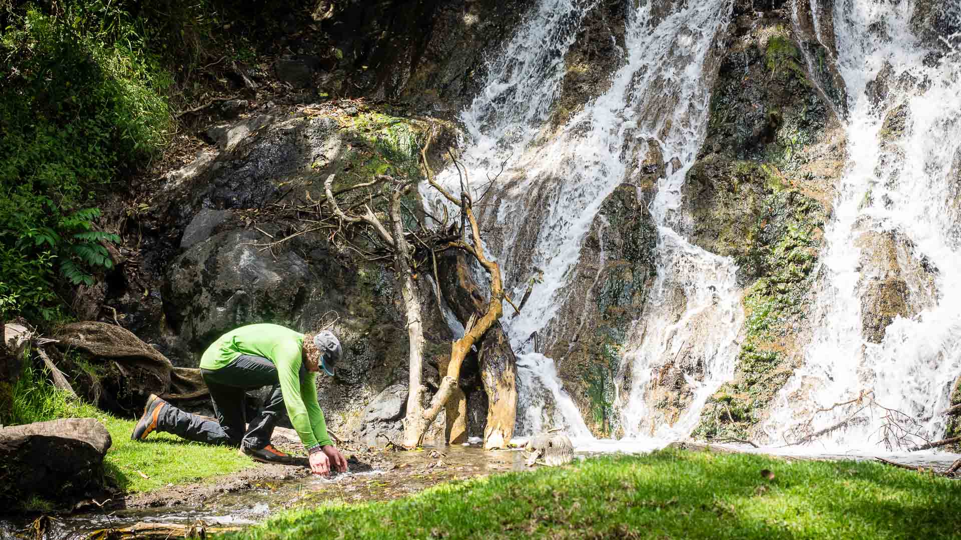 Trekker kauert an einem Rinnsal vor bemooster Felswand mit mehrstufigem Wasserfall am Kilimandscharo.
