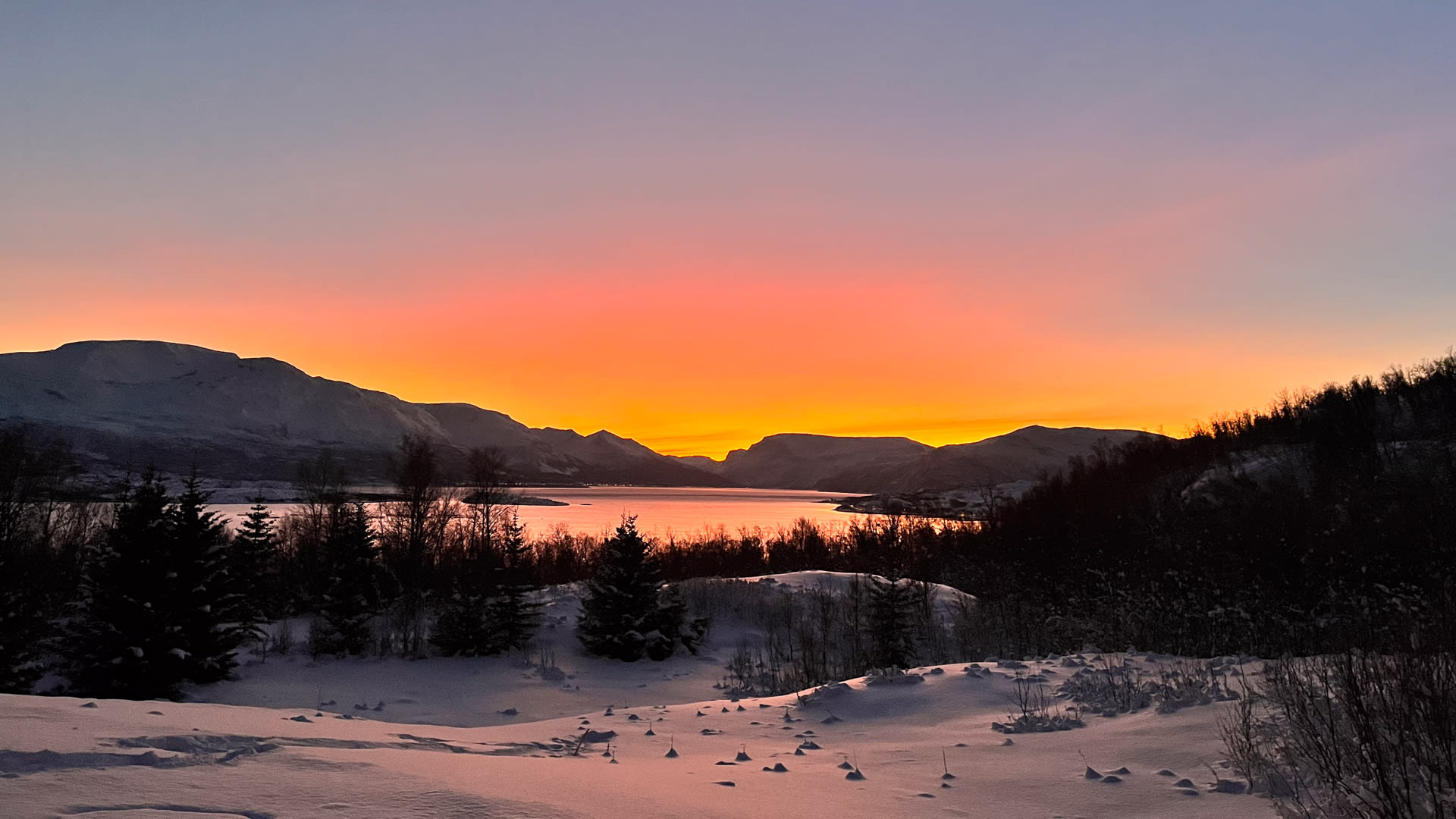 Winterwanderung in den Lyngen Alps: verschneite Fjordlandschaft mit Bergsilhouetten und orange-goldenem Abend- oder Morgenhimmel.