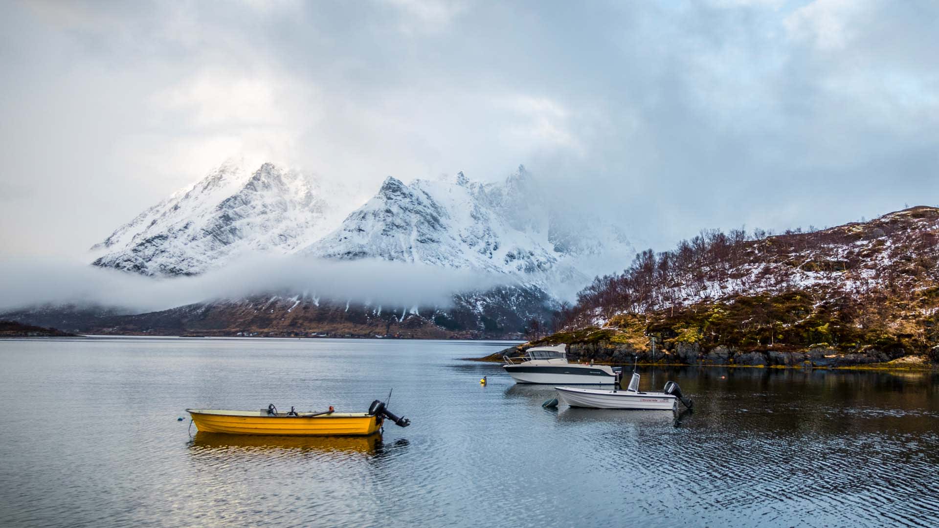 Winterwanderung Lofoten: vertäute Boote am ruhigen Fjord mit schneebedeckter Bergkette im Hintergrund.