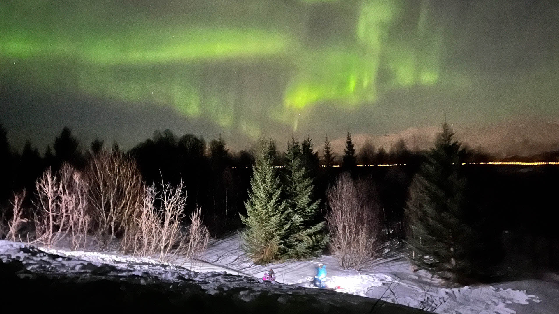 Zwei Winterwanderer sitzen im Schnee unter intensiv grünen Polarlichtern über einem Nadelwald in Norwegen bei Nacht.