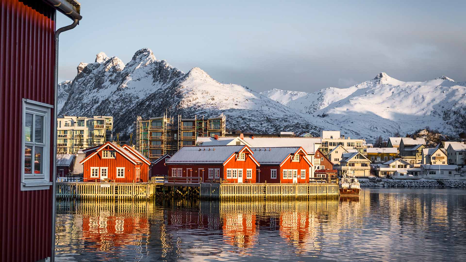 Rote Rorbu-Häuser in Svolvær spiegeln sich im Fjord, dahinter verschneite Berggipfel der Lofoten.