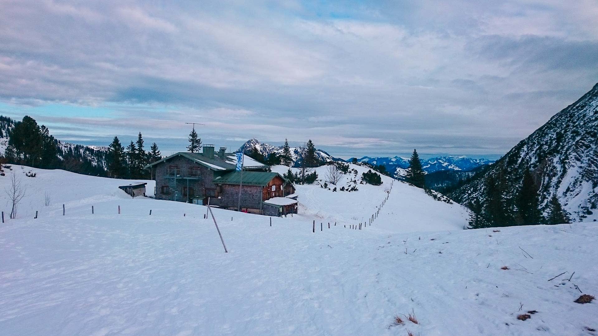 Winterwanderung zur Rotwandrunde am Spitzingsee: Taubensteinhaus mit grünem Blechdach auf verschneitem Hang mit Bergkulisse.