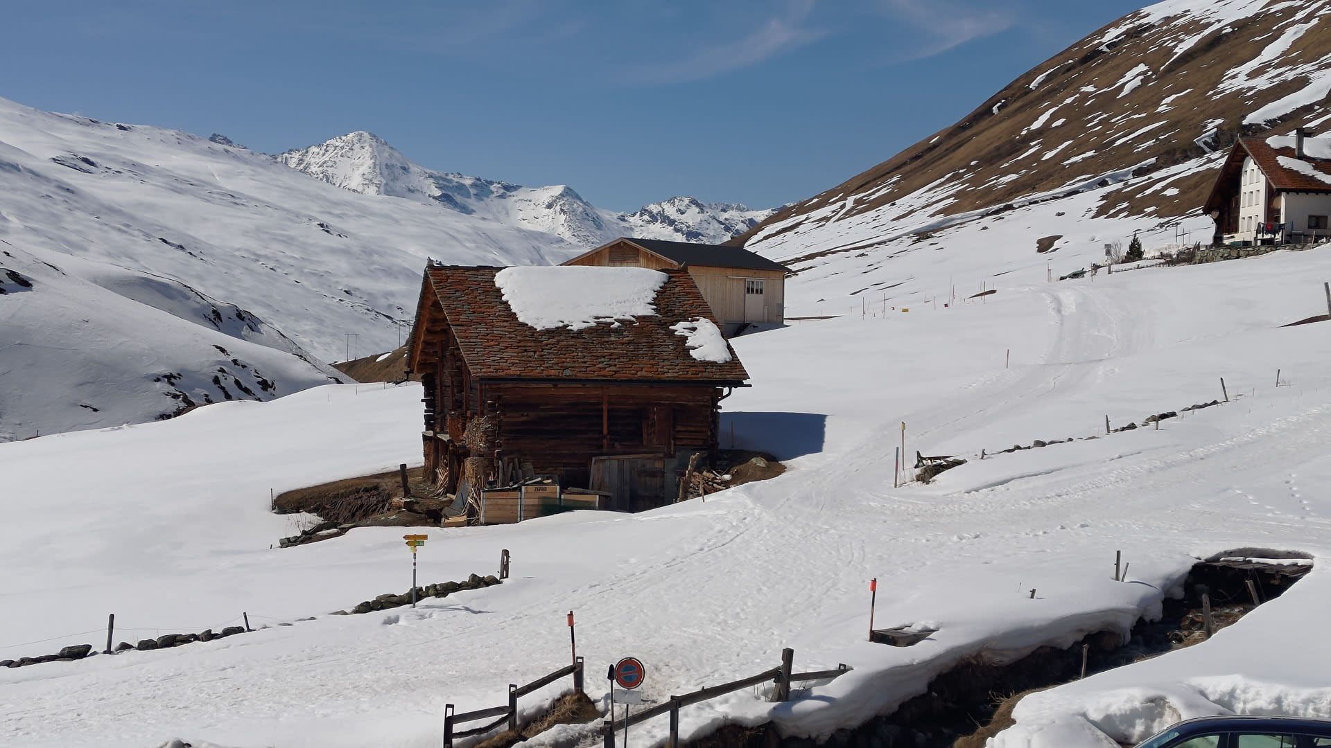 Winterwanderung im Averstal: verschneiter Weg mit Wegweiserschild, altem Steingebäude und vergletschertem Gipfel im Hintergrund.