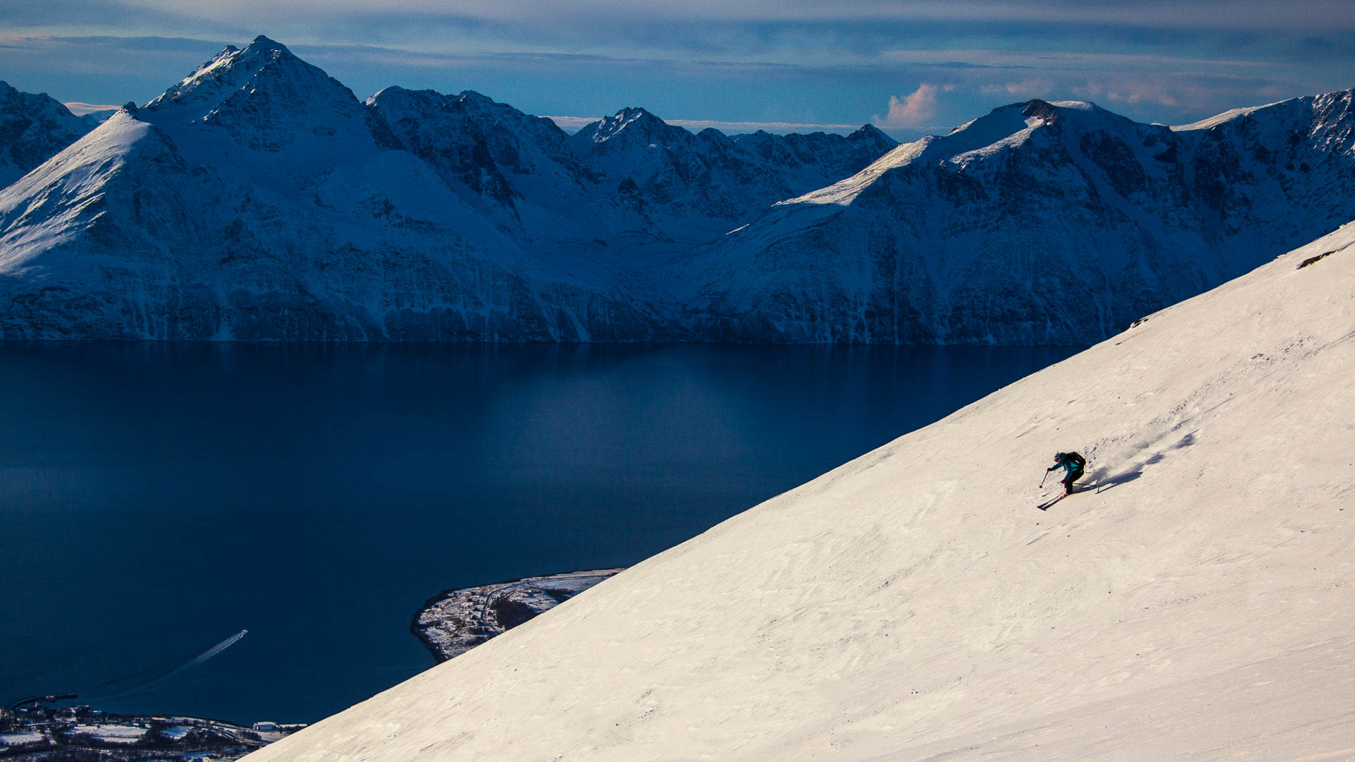 Skifahrer in blauer Jacke bei der Skitour in den Lyngen Alps fährt steilen Hang ab, im Hintergrund dunkelblauer Fjord