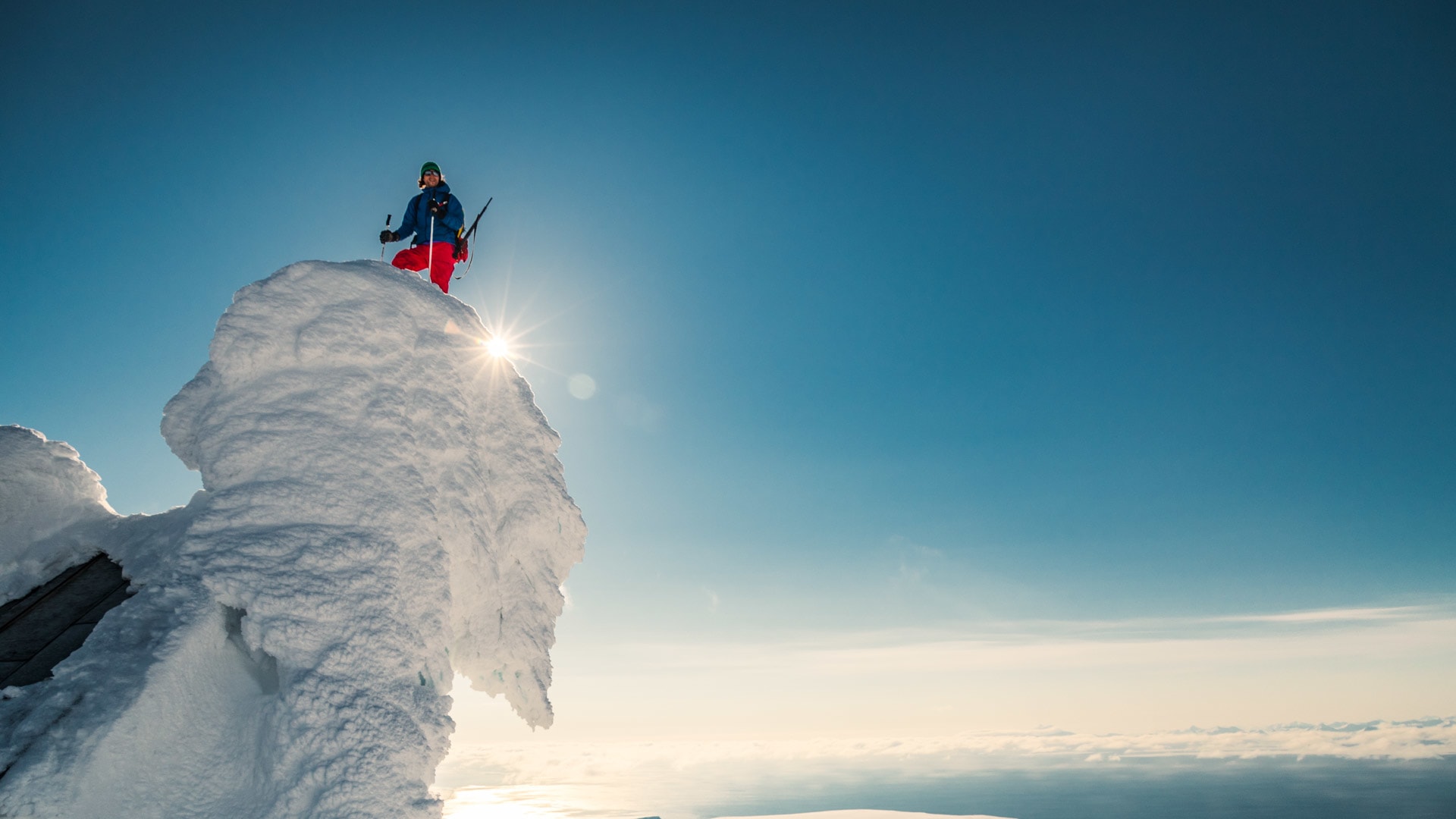 Ein Skitourengeher steht mit Skistöcken auf einem schneebedeckten Felsturm in Spitzbergen vor tiefblauem Himmel.