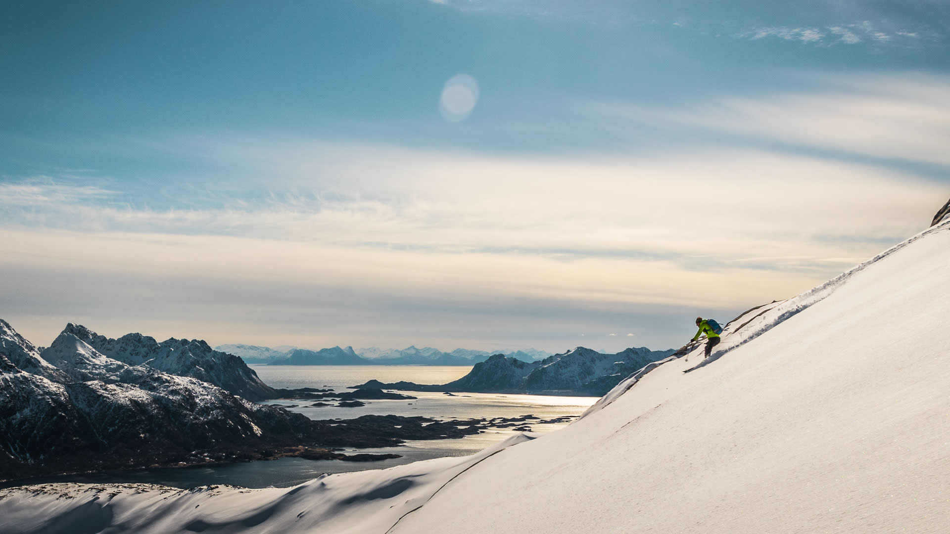 Skitourengeher in Abfahrtshaltung auf steilem Hang mit Fjord und Schären im Hintergrund auf den Lofoten.