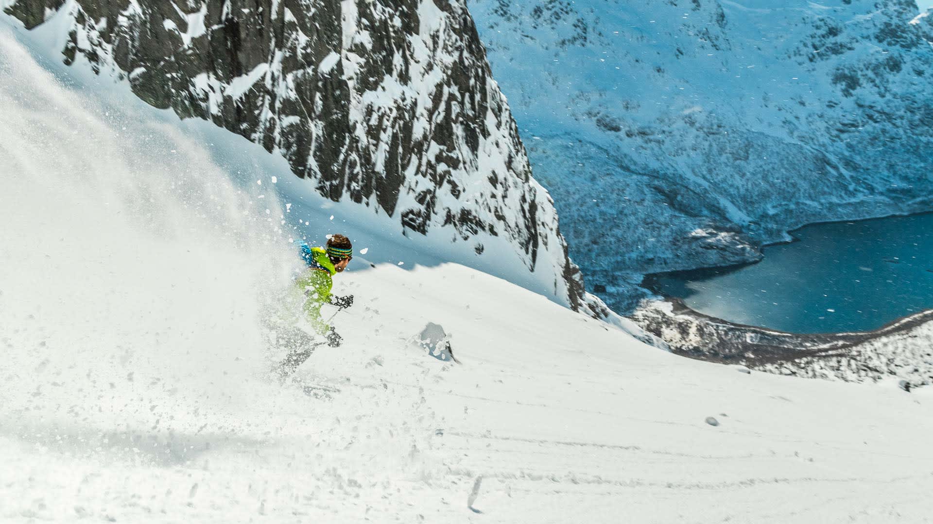 Skitourengeher fährt in gelbgrüner Jacke einen steilen Pulverschneehang mit Fjordblick auf den Lofoten ab.