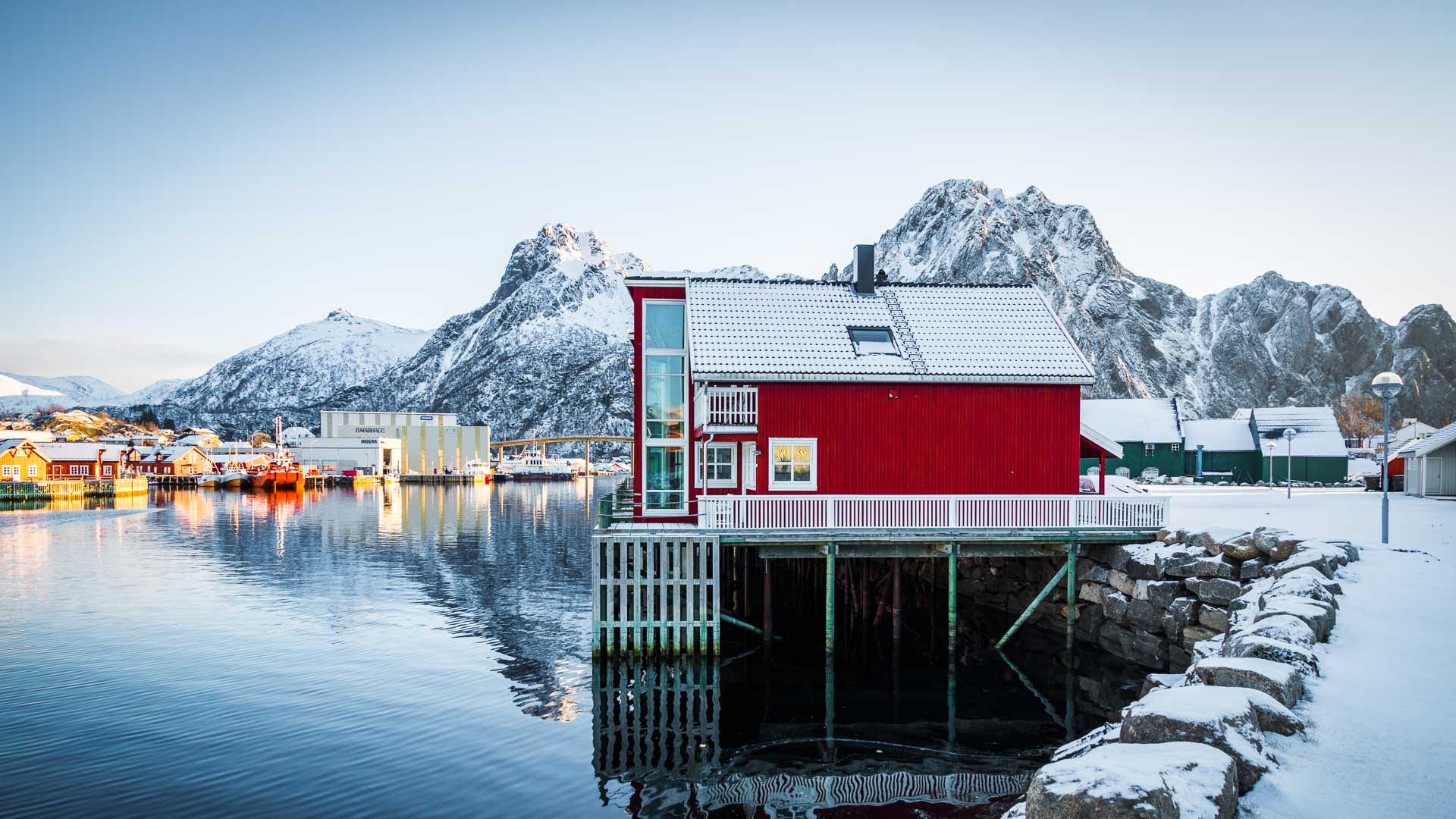 Skitour Lofoten: rotes Rorbu-Holzgebäude auf Pfählen über ruhigem Fjord, dahinter schneebedeckte Felsgrate und Bergmassive