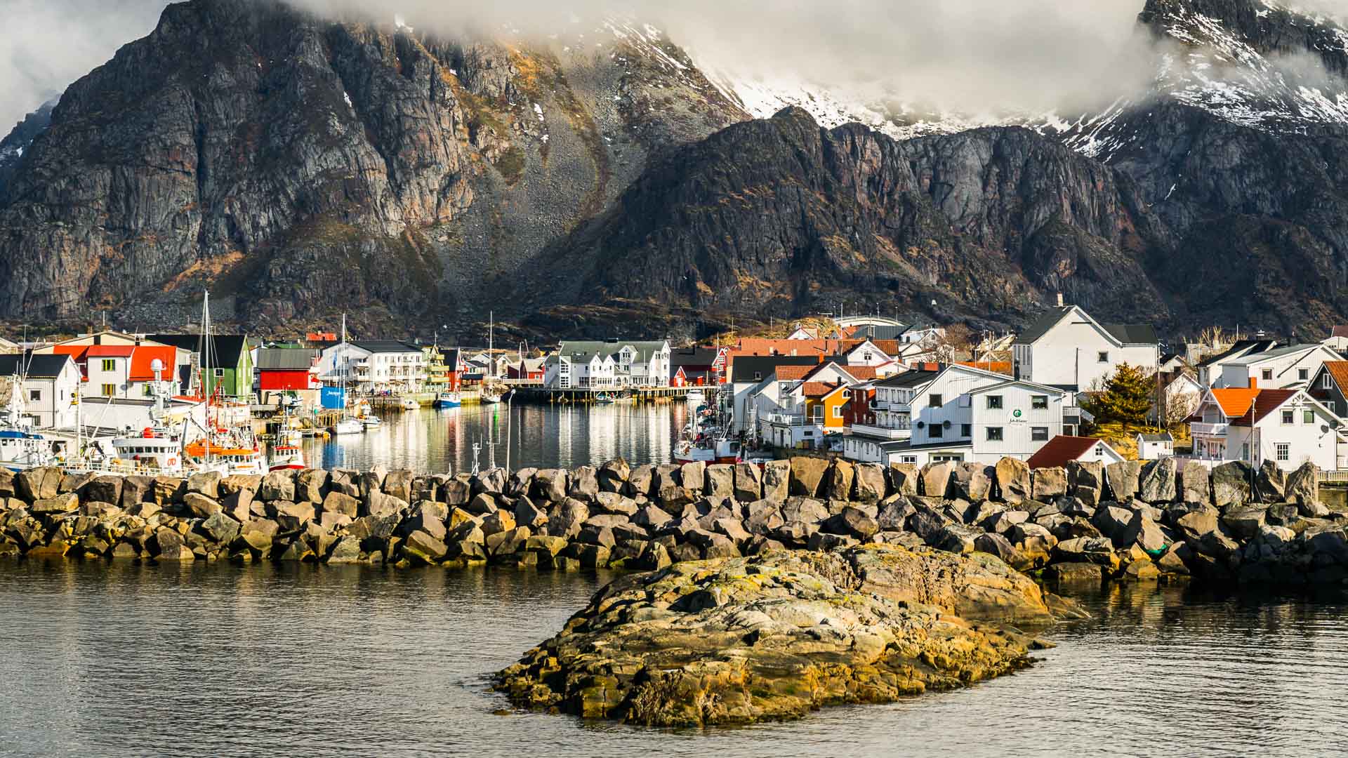 Blick auf den Fischerort Henningsvær auf den Lofoten mit Hafen, bunten Holzhäusern und schneebedeckter Felswand.