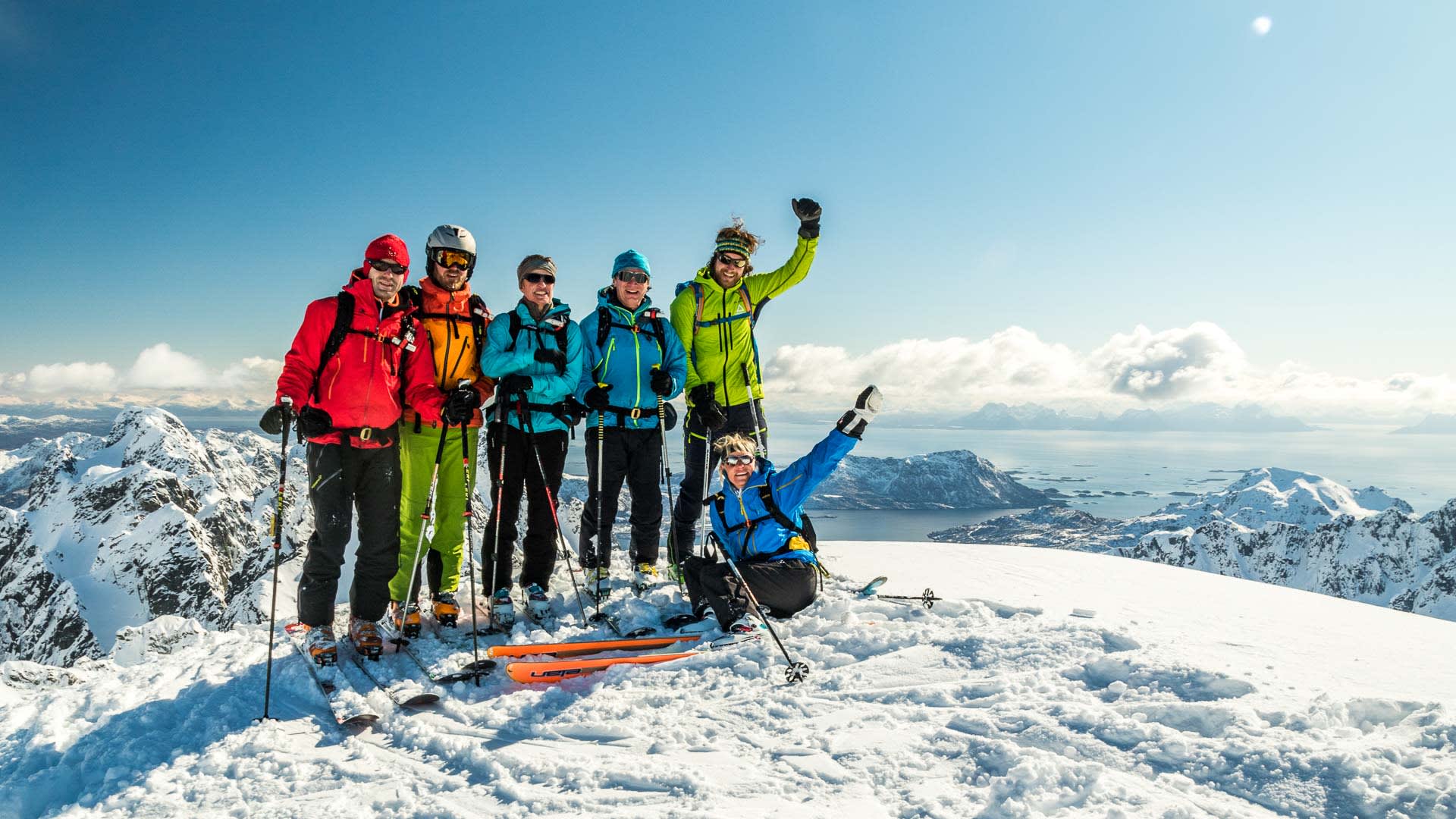 Sechs Skitourengeher posieren auf einem Gipfel der Lofoten mit Fjord- und Bergpanorama im Hintergrund.
