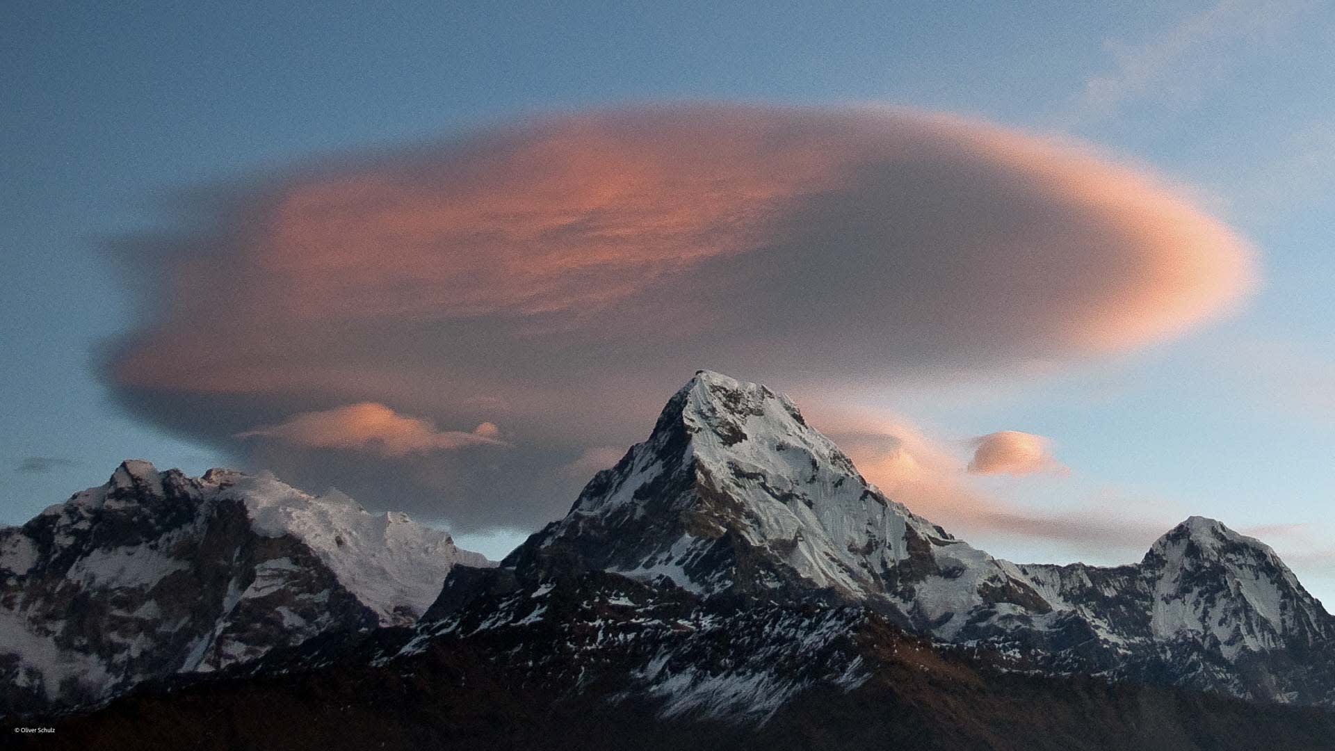 Trekking in hochalpinen Bergen: spitzer Gipfel mit Schnee und lentikularer Wolke in orange-rosa Farbtönen.