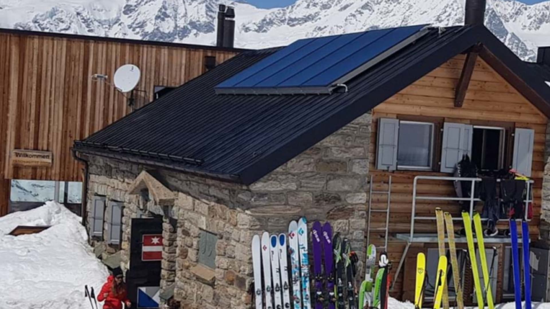 Zahlreiche bunte Tourenski stehen vor der Täschhütte im Schnee, zwei Personen sitzen daneben.