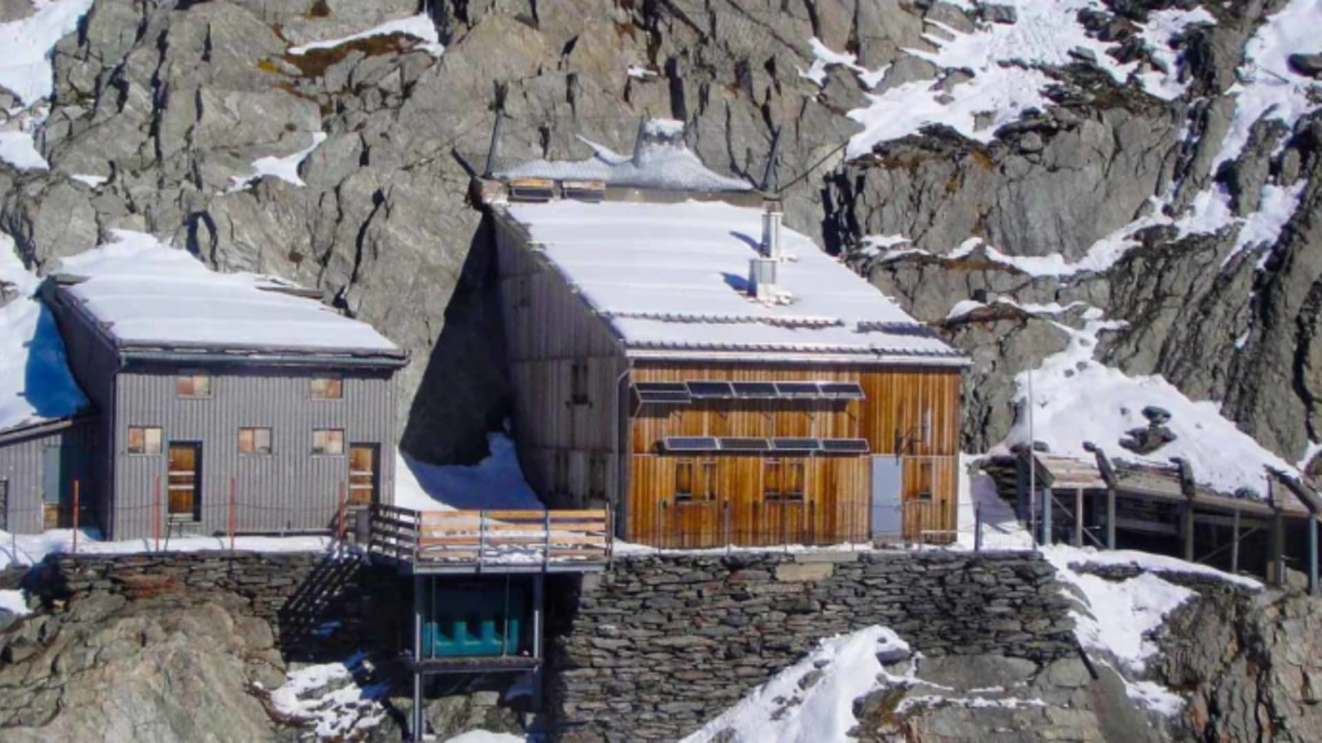 Luftaufnahme der Oberaarjochhütte mit grauen und orangefarbenen Gebäudeteilen auf verschneitem Felsgelände.