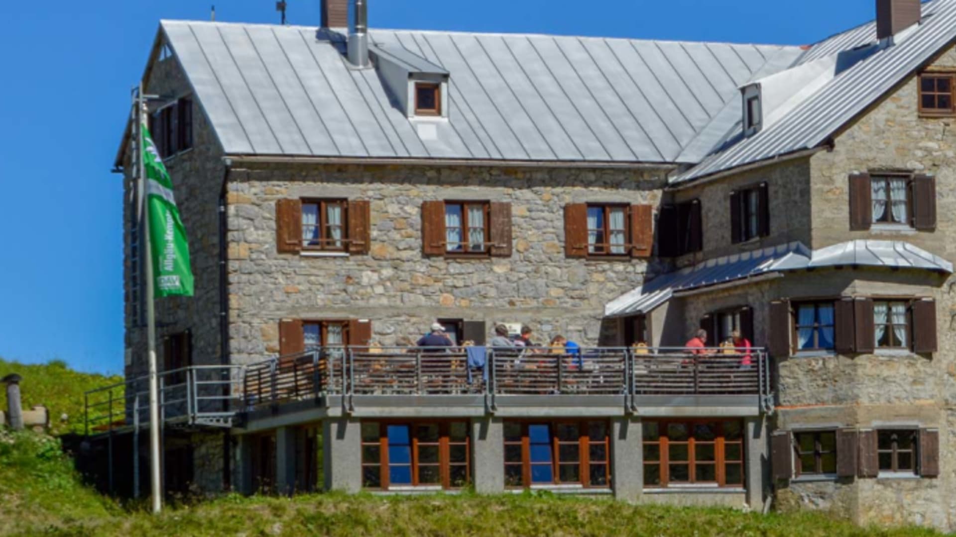 Außenansicht der Rappenseehütte mit Holzterrasse vor einer steilen Felswand in alpiner Umgebung.