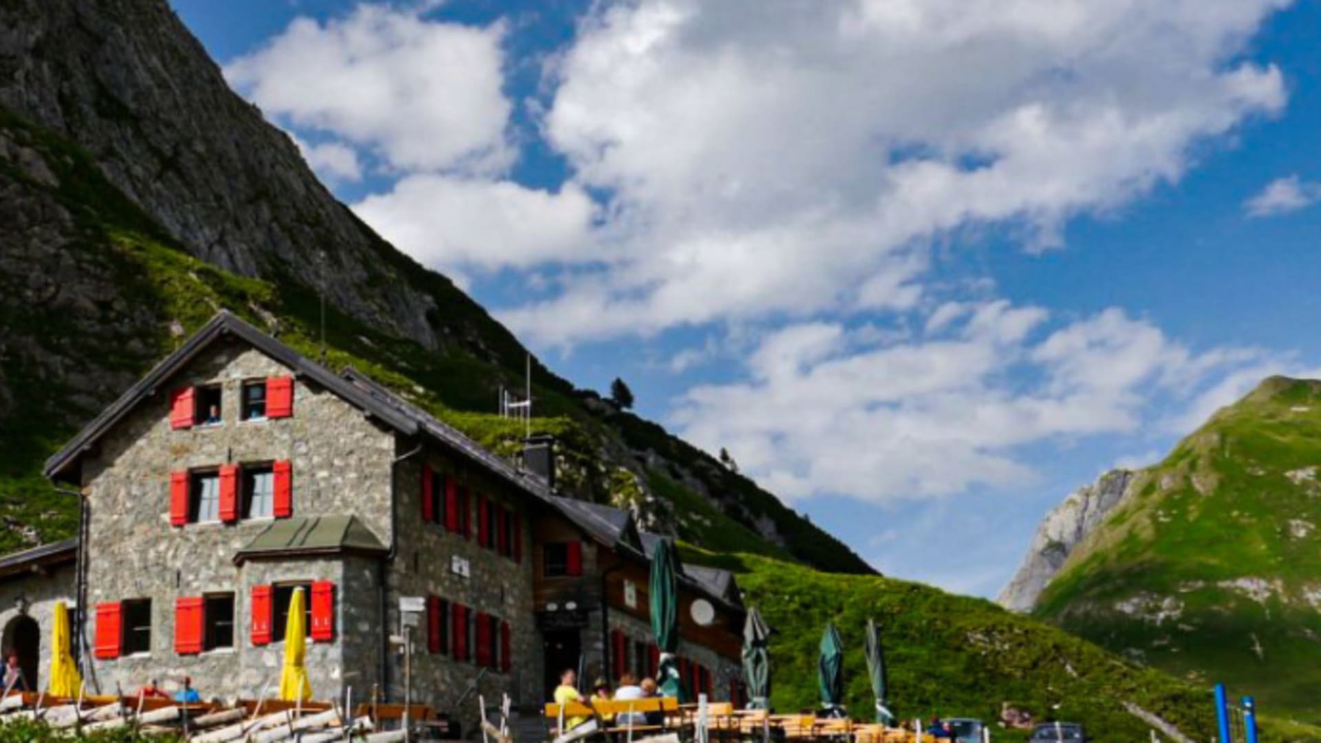 Die Ravensburger Hütte steht auf einer grünen Wiese mit großer Holzterrasse und gelben Sonnenschirmen.
