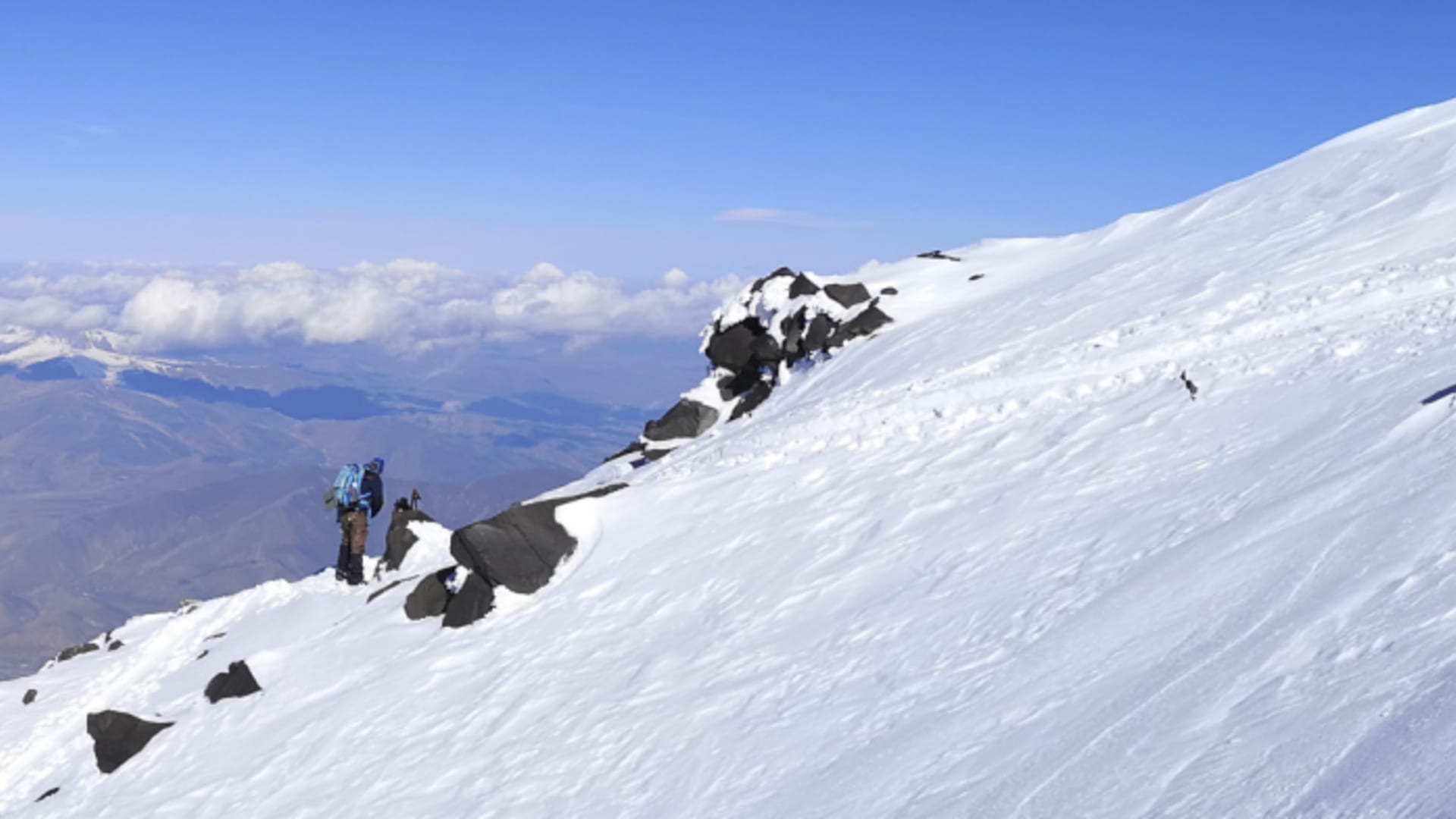 Zwei Höhenbergsteiger stehen auf einem Felsvorsprung am Schneehang des Ararat mit Blick ins weite Tal.