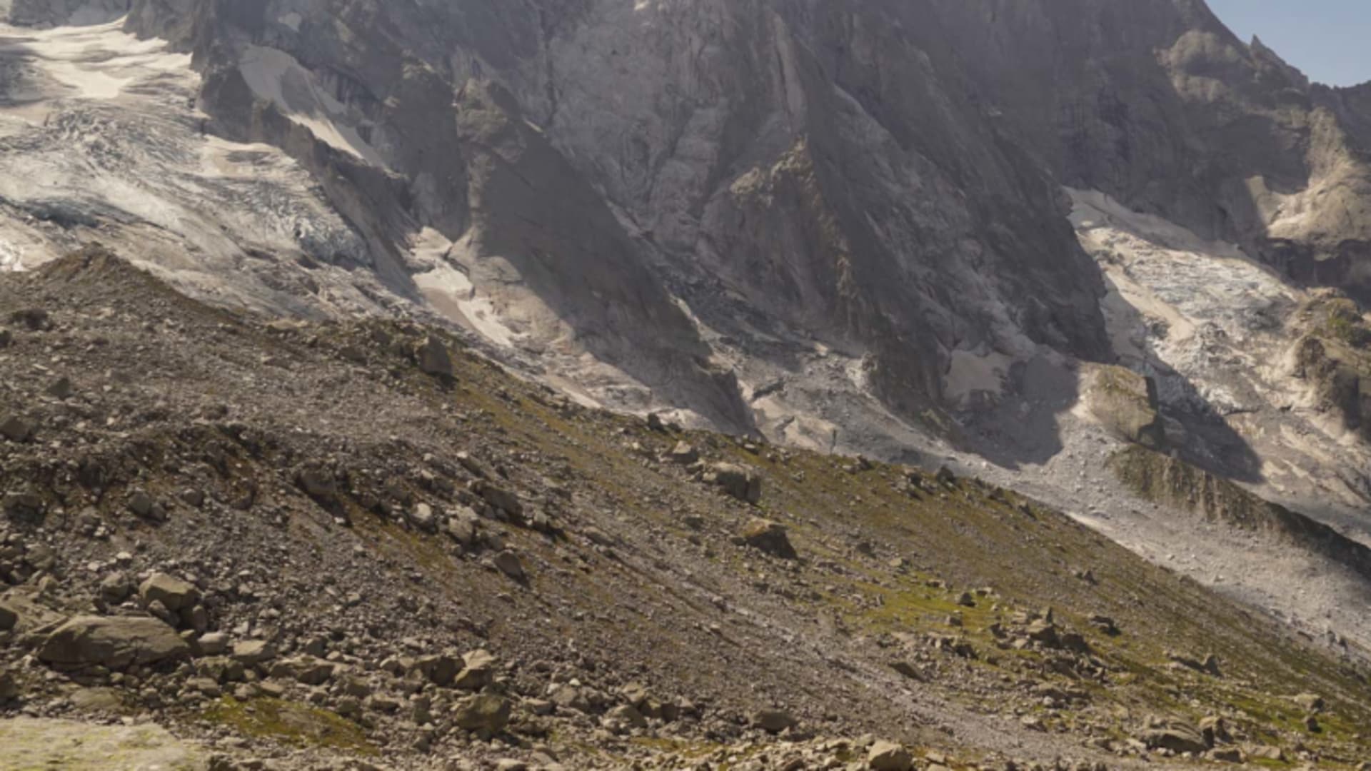 Bergwanderer mit Rucksack steigt auf grasig-steinigem Hang im Bergell mit Gletscherblick auf.