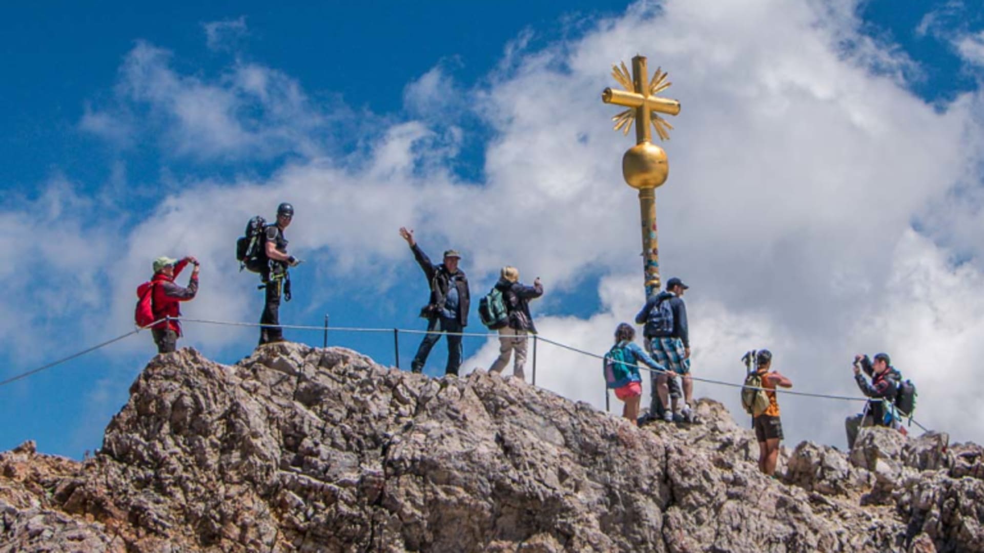 Bergwanderung auf die Zugspitze: sieben Bergwanderer am goldenen Gipfelkreuz auf dem felsigen Grat.