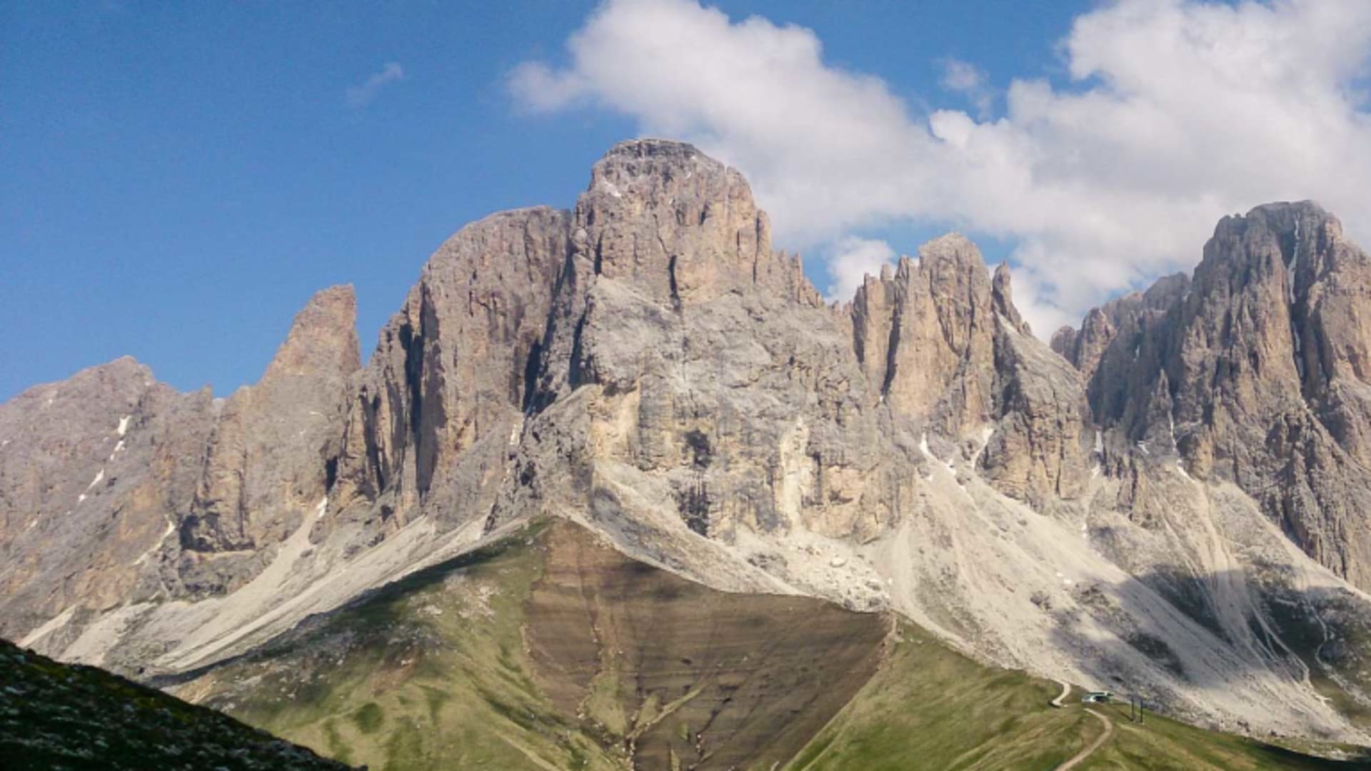 Bergwanderung am Sella Pass: Kalkfels-Massiv mit senkrechten Türmen, Schrofenhängen, Almwiesen und geschwungenem Fahrweg im Sommer.