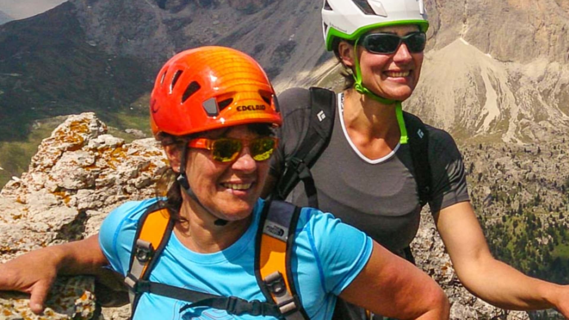 Zwei Klettersteiggeher mit Helm rasten auf felsbewachsenem Grat, im Hintergrund Dolomiten-Türme bei Cortina.