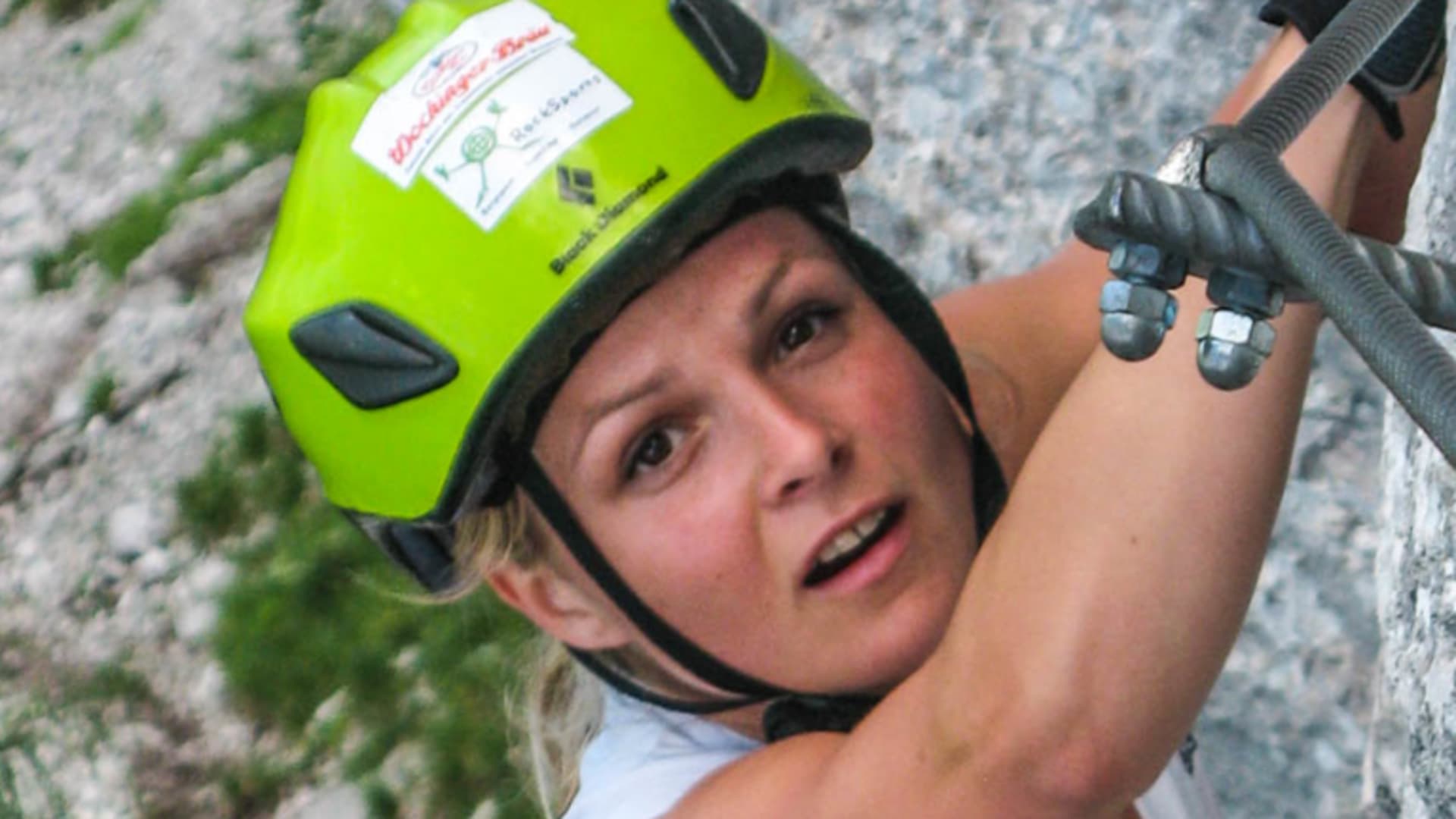 Klettersteiggeherin greift an Stahlbügel einer senkrechten Kalkfelsenwand am Staufen, gelber Helm.