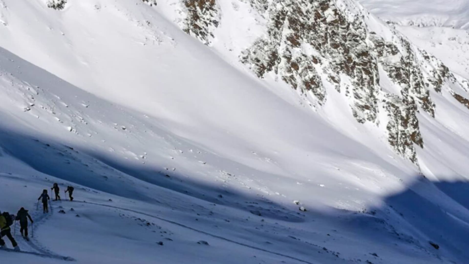 Sechs Skitourengeher steigen in Einzelreihe mit Fellen einen verschneiten Hang im Vinschgau auf.