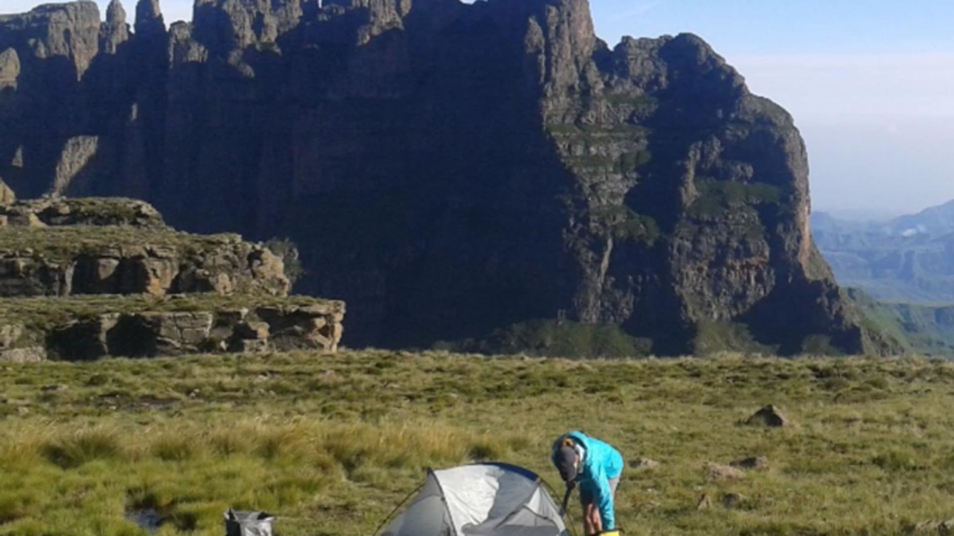 Trekker baut Kuppelzelt auf dem Hochplateau der Drakensberge auf, großer Rucksack liegt daneben, steile Felswand im Hintergrund.