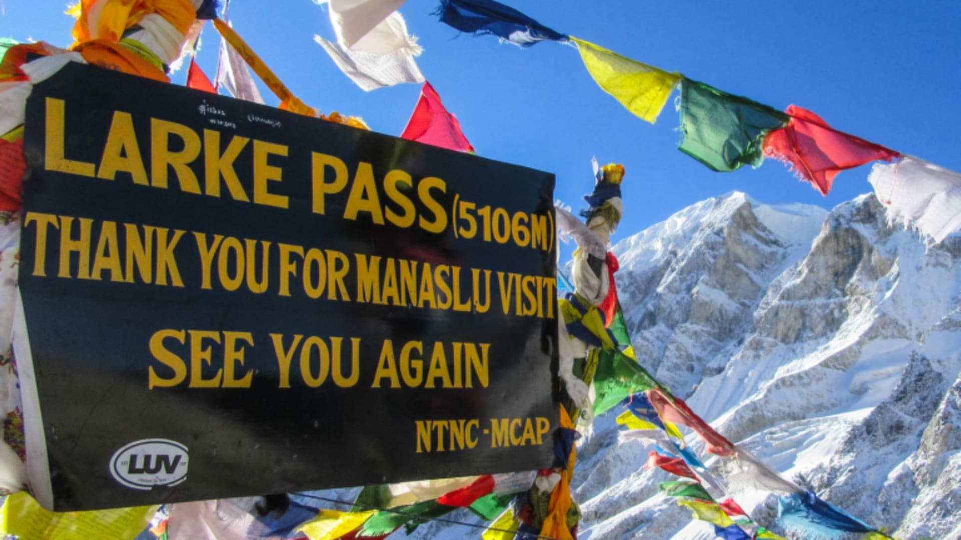 Trekking am Larkya Pass (5106 m) mit Gipfelschild, bunten Gebetsfahnen und verschneitem Hochgebirgsgipfel im Hintergrund.