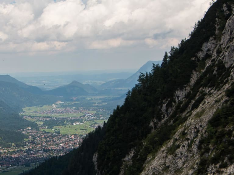 Panoramablick vom Kreuzeckhaus hinab ins Tal nach Garmisch-Partenkirchen mit Siedlung und bewaldeten Berghängen.