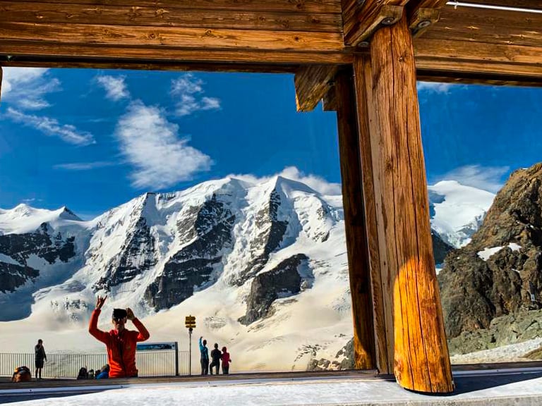 Blick aus dem Berghaus Diavolezza durch die Holzkonstruktion auf Besucher und vergletscherte Berggipfel.