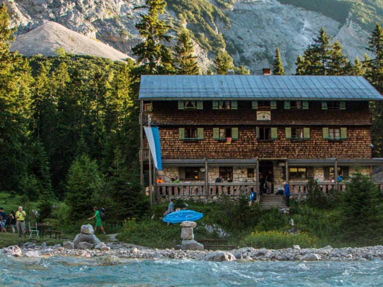 Die Reintalangerhütte im warmen Abendlicht am steinigen Gebirgsbach mit Wanderern auf der Terrasse.
