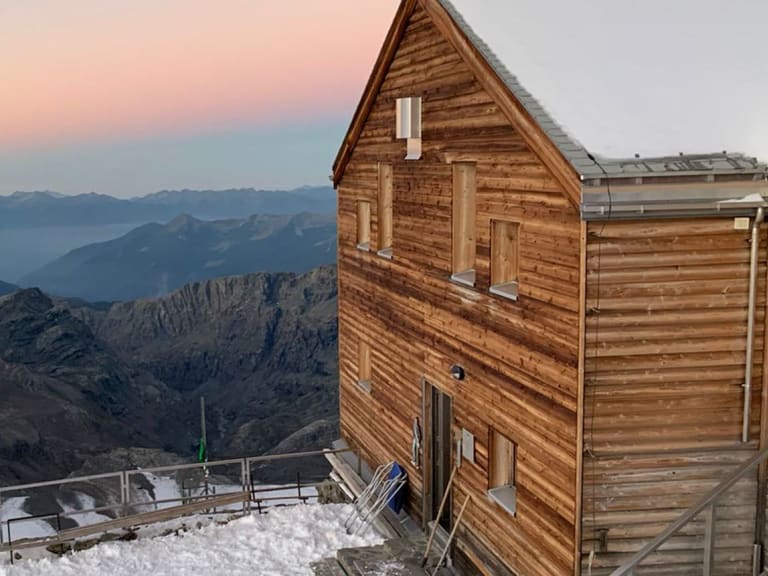 Außenansicht der Marco e Rosa Hütte in Holzbauweise mit beleuchteten Fenstern und weitem Blick über verschneite Gipfel d