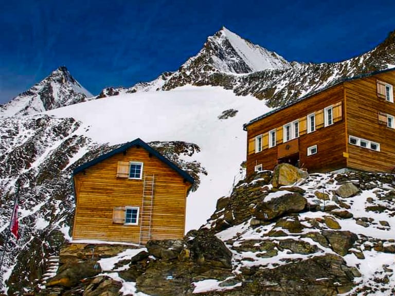 Die Mischabelhütte besteht aus zwei Holzgebäuden auf felsigem Terrain vor schneebedeckten Gipfeln der Walliser Alpen.