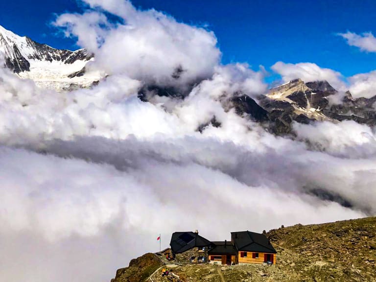 Panoramablick auf die Domhütte über dichter Wolkendecke mit schneebedeckten Gipfeln im Hintergrund.