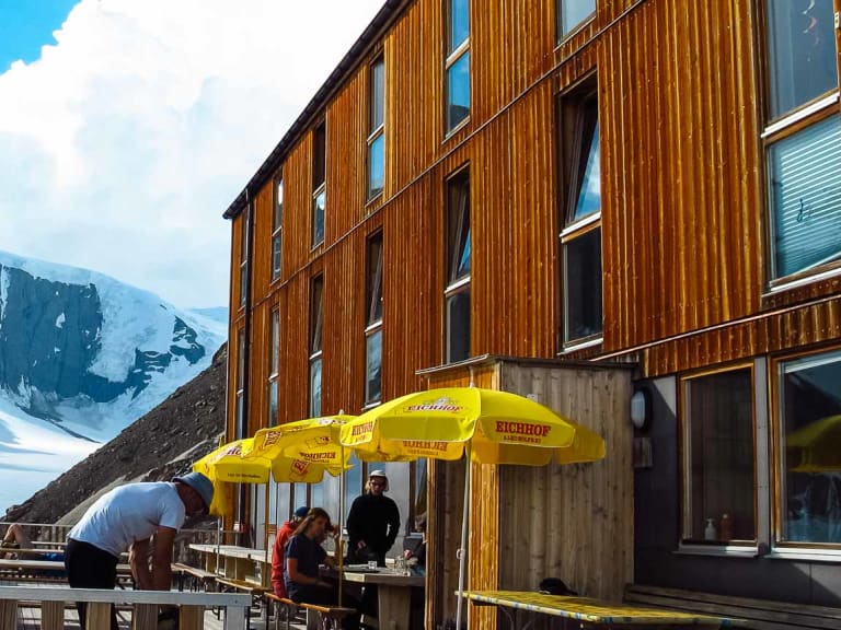 Gäste entspannen unter gelben Sonnenschirmen auf der Holzterrasse der Finsteraarhornhütte vor Gletscherkulisse.