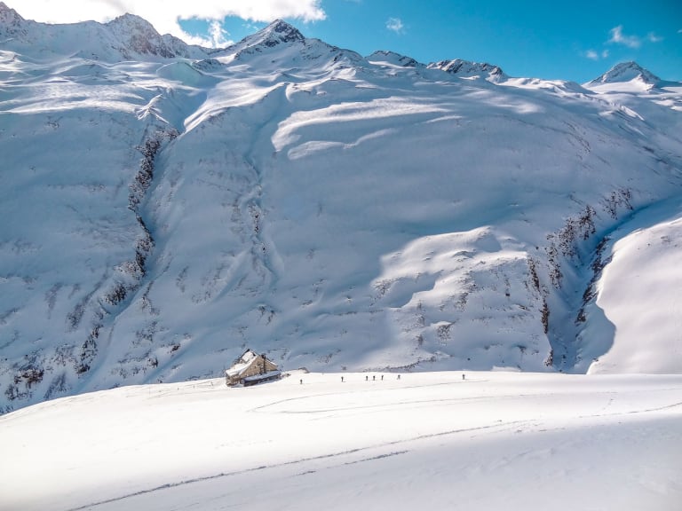 Winteraufnahme der Vernagthütte mit Skifahrern auf verschneiter Fläche vor Bergkulisse der Ötztaler Alpen.