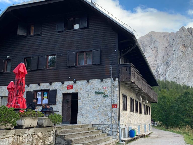 Berghaus Tičarjev dom na Vršiču vor imposanter Kalksteinwand mit Terrasse und Wegweiser.