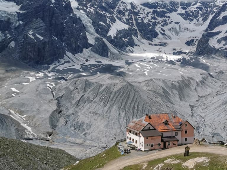 Das Rifugio Casati steht auf grüner Almwiese vor ausgedehntem Geröllfeld und schneebedeckter Gipfelkette mit Gletscher.