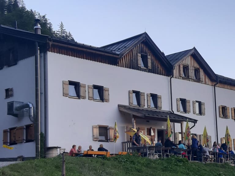Außenansicht der Gaudeamushütte mit Terrasse und Gästen auf grüner Almwiese im Wilden Kaiser.
