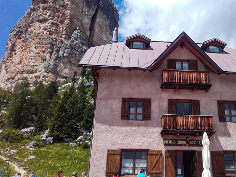 Wanderer vor dem Rifugio Cinque Torri, das unterhalb einer steilen Kalksteinwand in den Dolomiten auf Almwiesen steht.