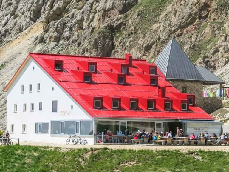 Bergwanderer mit Rucksack wandert zur Tierser Alpl Hütte mit weißer Fassade und rotem Dach.